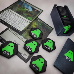 May include: A selection of Magic: The Gathering game accessories. Includes green and black Ooze creature tokens, a card holder, and game cards. The tokens feature a green ghost design with 0/0 text. The cards are labeled "Slime Against Humanity" and "Token Creature - Ooze".