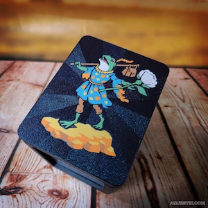 以下が含まれることがあります： 黒いケースに、カバンと白いバラを持ったカエルのキャラクターのカラフルなイラストが描かれています。カエルはオレンジ色の水玉模様の青い衣装を着ています。ケースは木製の表面に置かれています。