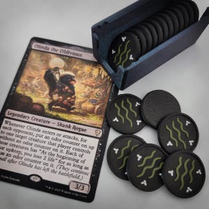 May include: A Magic: The Gathering trading card titled Olinda the Oblivious featuring a skunk illustration, accompanied by a dark blue plastic storage box containing several black circular tokens with green wavy lines and white accents.