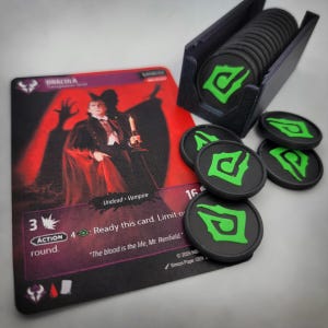 May include: A Dracula character card with red and black artwork, featuring the text "Dracula" and "The blood is the life, Mr. Renfield." Next to the card is a black plastic holder containing a stack of black circular tokens with green symbols.