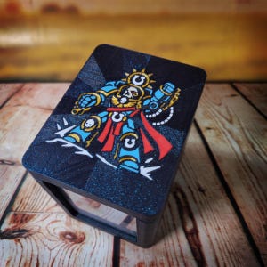 May include: A rectangular black storage box featuring a glittery finish and a colorful graphic on the lid. The graphic depicts a stylized armored warrior in blue, red, and gold tones, complete with a skull emblem and a prominent U-shaped symbol.