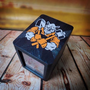 May include: A black rectangular box featuring a graphic on the top lid. The illustration depicts a rhinoceros wearing orange clothing and holding a shovel and a hammer. The box has a textured finish and a clear front panel showing the interior space.