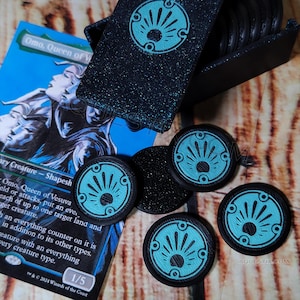 May include: Five black and blue plastic tokens with a sunburst design. The tokens are in a box with a blue glitter finish. The box is open and the tokens are spilling out.