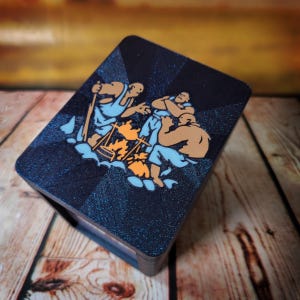 May include: A rectangular box with a dark blue glitter finish featuring a graphic illustration of three figures gathered around a campfire. The figures are depicted in light blue and tan tones, with orange flames visible in the center of the scene.