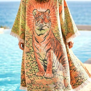 Op de afbeelding: Een lange, aan de voorzijde open kimono-stijl badjas met een grote, gedetailleerde afbeelding van een tijger te midden van gebladerte. Het kledingstuk is voornamelijk oranje, geel en groen, met een zonnestraalpatroon en witte franjes aan de zoom.