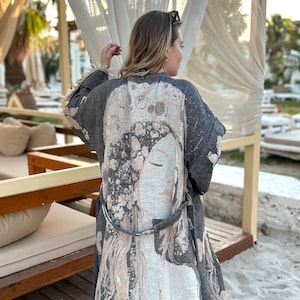 May include: A gray kimono-style robe with a unique print featuring a woman's face and abstract patterns in shades of beige and gray. The robe has a belt, fringe trim, and is worn outdoors. The background includes beach scenery.