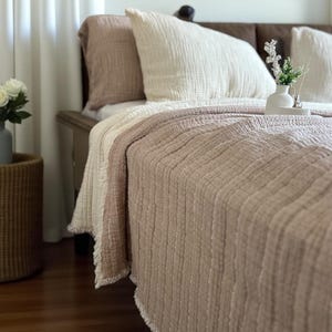 May include: A bedroom scene featuring a bed with a light brown textured quilt and white pillows. A woven basket holds a vase of white roses. White curtains and a built-in shelf with decor complete the cozy, neutral-toned space.
