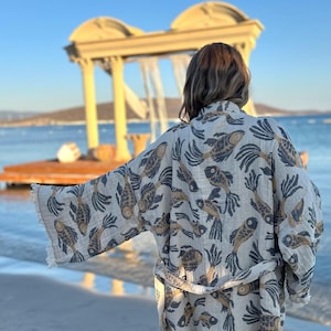 May include: A beige robe with a fish pattern in shades of brown and navy blue. The robe has a matching belt and fringe detailing on the sleeves. The image is taken on a beach with a view of the ocean and a structure in the background.