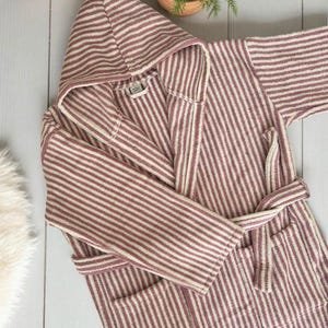 May include: A hooded, striped robe in shades of burgundy and cream. The robe features a matching belt, pockets, and a soft, textured fabric. The robe is designed for comfort and relaxation.
