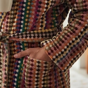 May include: A patterned dressing gown in a variety of colours, including brown, yellow, pink, and green, with a belt. A person is holding a white mug. The robe has pockets. The person is wearing the robe.