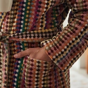 May include: A multi-colored patterned robe with a belt, held by a person. The robe features a mix of colors including brown, yellow, pink, and green. The person is holding a white mug. The robe has pockets.