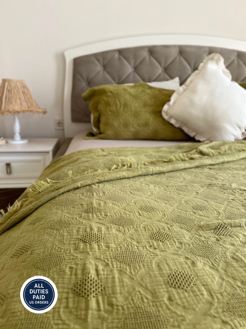 K&ouml;nnte beinhalten: Ein Bett mit getuftetem Kopfteil, olivgr&uuml;nen Kissen und einer passenden gemusterten Decke. Auf der linken Seite befindet sich ein wei&szlig;er Nachttisch mit einer Lampe mit gewebtem Schirm. Die Decke hat ein strukturiertes, florales Design.