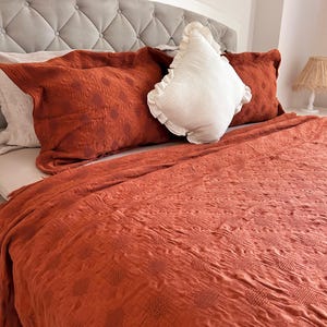 K&ouml;nnte beinhalten: Ein Bett mit einem grau getufteten Kopfteil und rostfarbener Bettw&auml;sche. Zwei rostfarbene Kissen mit einem gepunkteten Muster und ein wei&szlig;es Kissen mit R&uuml;schen liegen auf dem Bett. Eine rostfarbene Decke bedeckt das Bett.