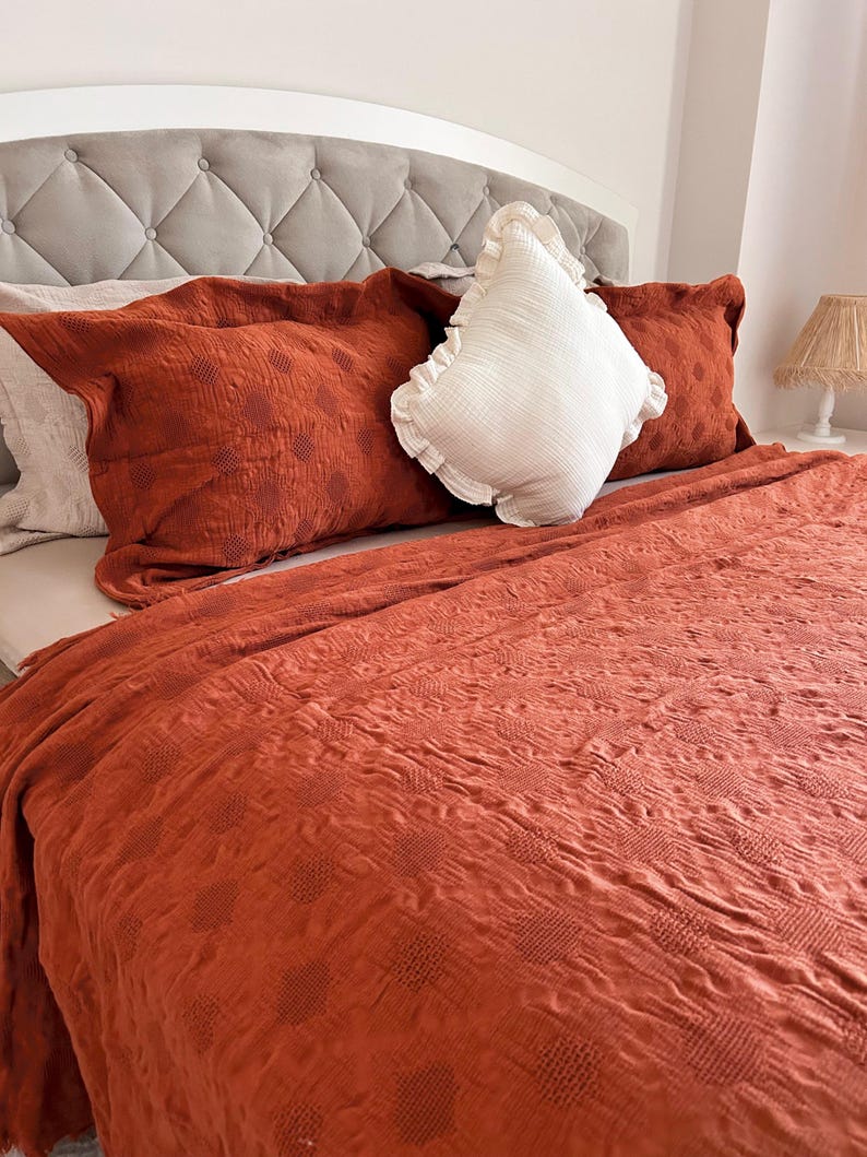 K&ouml;nnte beinhalten: Ein Bett mit einem grau getufteten Kopfteil und rostfarbener Bettw&auml;sche. Zwei rostfarbene Kissen mit einem gepunkteten Muster und ein wei&szlig;es Kissen mit R&uuml;schen liegen auf dem Bett. Eine rostfarbene Decke bedeckt das Bett.