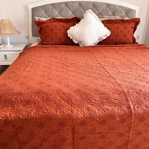 K&ouml;nnte beinhalten: Ein Bett mit wei&szlig;em Kopfteil und einem grau gepolsterten Paneel. Das Bett ist mit einer orange-roten, strukturierten Steppdecke und passenden Kissen bedeckt. Ein wei&szlig;es, ger&uuml;schtes Kissen liegt obenauf. Ein wei&szlig;er Nachttisch mit Lampe befindet sich links.