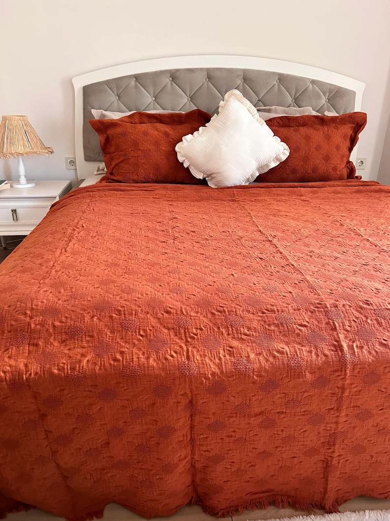 K&ouml;nnte beinhalten: Ein Bett mit wei&szlig;em Kopfteil und einem grau gepolsterten Paneel. Das Bett ist mit einer orange-roten, strukturierten Steppdecke und passenden Kissen bedeckt. Ein wei&szlig;es, ger&uuml;schtes Kissen liegt obenauf. Ein wei&szlig;er Nachttisch mit Lampe befindet sich links.