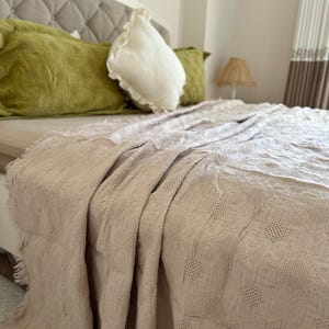 K&ouml;nnte beinhalten: Beige Tagesdecke mit strukturiertem, gemustertem Design, drapiert &uuml;ber einem Bett. Das Bett ist mit gr&uuml;nen und wei&szlig;en Kissen ausgestattet. Im Hintergrund ist ein wei&szlig;es Kopfteil zu sehen. Der Raum hat eine neutrale Farbpalette.