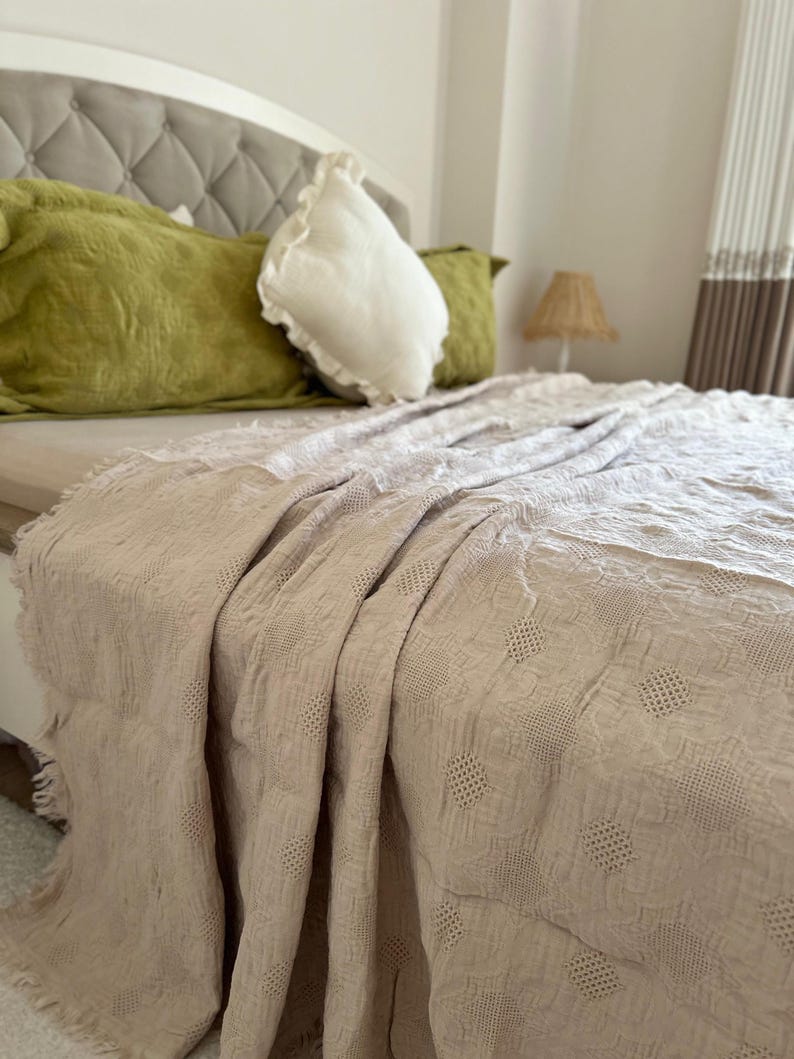 K&ouml;nnte beinhalten: Beige Tagesdecke mit strukturiertem, gemustertem Design, drapiert &uuml;ber einem Bett. Das Bett ist mit gr&uuml;nen und wei&szlig;en Kissen ausgestattet. Im Hintergrund ist ein wei&szlig;es Kopfteil zu sehen. Der Raum hat eine neutrale Farbpalette.