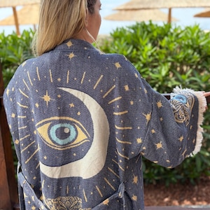 May include: A dark blue woven robe featuring a large cream-colored crescent moon and a central eye graphic with light blue iris details. The fabric is decorated with gold star patterns and includes a matching tie belt and fringed sleeve cuffs.