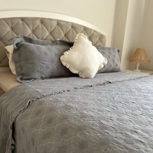 K&ouml;nnte beinhalten: Ein Bett mit einem grau getufteten Kopfteil und einer grauen Tagesdecke. Das Bett ist mit grauen Kissen und einem wei&szlig;en Kissen mit R&uuml;schen ausgestattet. Eine kleine Lampe mit gewebtem Schirm steht auf dem Nachttisch.