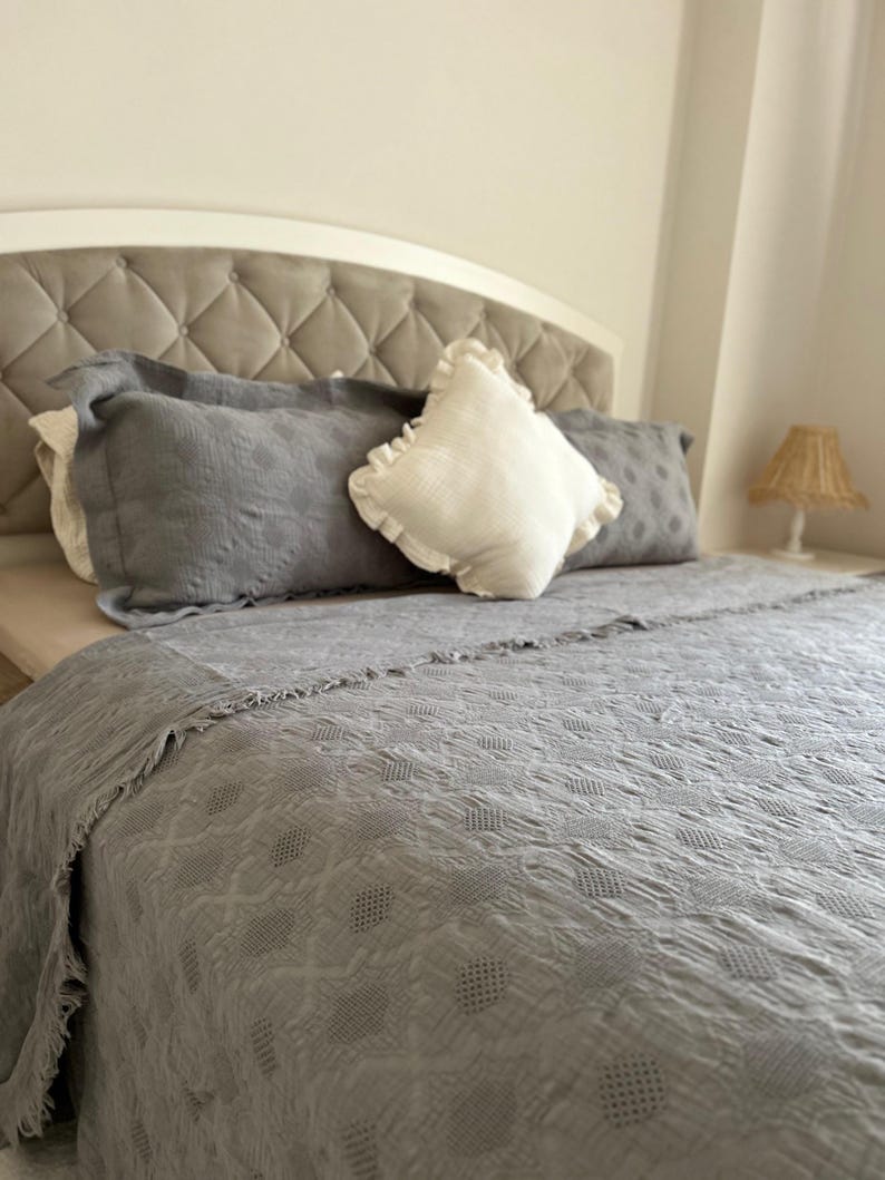 K&ouml;nnte beinhalten: Ein Bett mit einem grau getufteten Kopfteil und einer grauen Tagesdecke. Das Bett ist mit grauen Kissen und einem wei&szlig;en Kissen mit R&uuml;schen ausgestattet. Eine kleine Lampe mit gewebtem Schirm steht auf dem Nachttisch.
