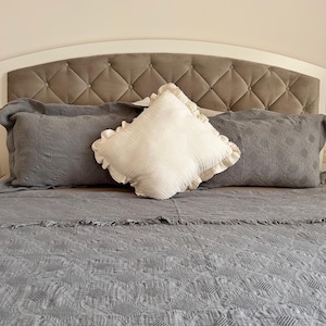 K&ouml;nnte beinhalten: Ein Bett mit einer grauen, gesteppten Tagesdecke, zwei grauen Kissen und einem wei&szlig;en, quadratischen Kissen mit R&uuml;schen. Das Kopfteil ist wei&szlig; mit einem rautenf&ouml;rmig getufteten grauen Mittelteil.