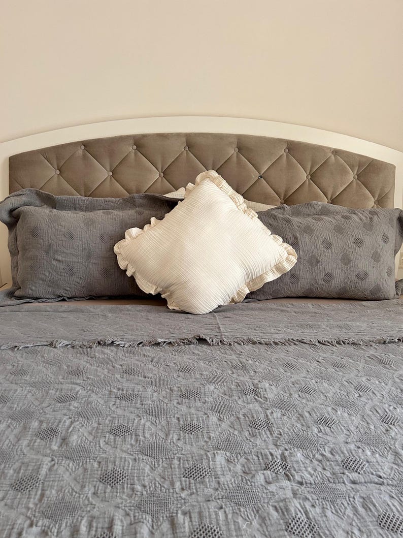 K&ouml;nnte beinhalten: Ein Bett mit einer grauen, gesteppten Tagesdecke, zwei grauen Kissen und einem wei&szlig;en, quadratischen Kissen mit R&uuml;schen. Das Kopfteil ist wei&szlig; mit einem rautenf&ouml;rmig getufteten grauen Mittelteil.