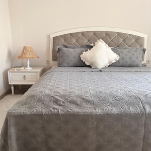 K&ouml;nnte beinhalten: Ein Bett mit einer grauen, gesteppten Tagesdecke und passenden Kissen. Das Kopfteil ist wei&szlig; mit einem grauen Stoff mit Rautenmuster. Ein wei&szlig;es, ger&uuml;schtes Kissen liegt auf dem Bett. Ein wei&szlig;er Nachttisch mit einer Lampe befindet sich links.