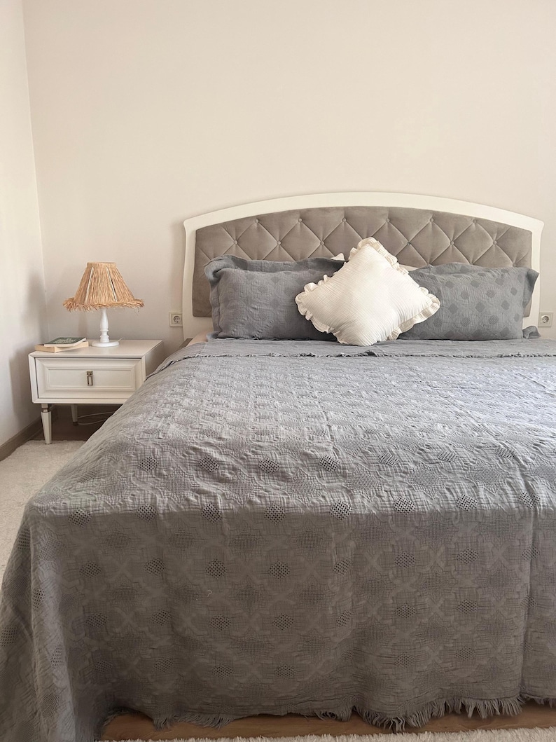 K&ouml;nnte beinhalten: Ein Bett mit einer grauen, gesteppten Tagesdecke und passenden Kissen. Das Kopfteil ist wei&szlig; mit einem grauen Stoff mit Rautenmuster. Ein wei&szlig;es, ger&uuml;schtes Kissen liegt auf dem Bett. Ein wei&szlig;er Nachttisch mit einer Lampe befindet sich links.