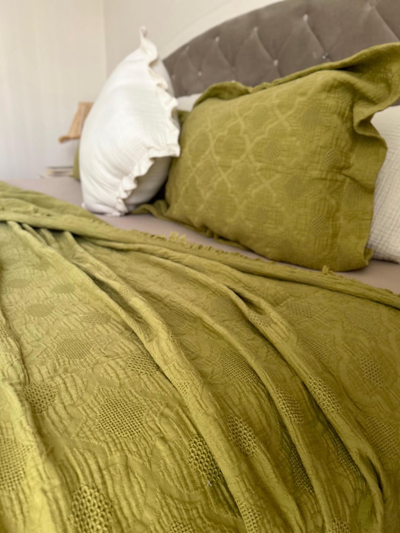 K&ouml;nnte beinhalten: Ein Bett mit olivgr&uuml;ner Bettw&auml;sche, darunter eine gemusterte Decke und dekorative Kissen. Ein wei&szlig;es Kissen mit R&uuml;schen ist ebenfalls zu sehen. Das Kopfteil ist in einem neutralen Grauton gehalten.