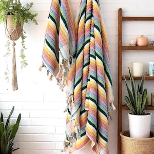 May include: Two colourful striped beach towels with tassels hanging on a white brick wall. The towels feature stripes of yellow, pink, green, and blue. A wooden ladder shelf and potted plants are in the background.