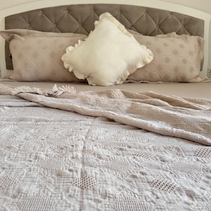 K&ouml;nnte beinhalten: Ein Bett mit einem getufteten Kopfteil, verziert mit beigen Kissen und einem dekorativen wei&szlig;en Kissen mit R&uuml;schenkanten. Das Bett ist mit einer beige, aufwendig gemusterten Steppdecke bedeckt, was eine gem&uuml;tliche Schlafzimmerumgebung schafft.