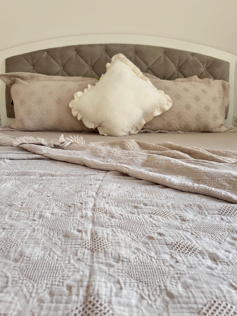 K&ouml;nnte beinhalten: Ein Bett mit einem getufteten Kopfteil, verziert mit beigen Kissen und einem dekorativen wei&szlig;en Kissen mit R&uuml;schenkanten. Das Bett ist mit einer beige, aufwendig gemusterten Steppdecke bedeckt, was eine gem&uuml;tliche Schlafzimmerumgebung schafft.