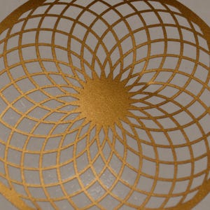 May include: A circular gold-colored cutout featuring a complex, repeating geometric pattern of intersecting curved lines that radiate from a solid center point, creating a symmetrical lattice design across the entire round surface.