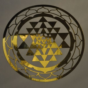 May include: A gold-colored, circular decorative piece featuring a Sri Yantra design. The intricate pattern includes interlocking triangles and a lotus flower border. The piece has a metallic sheen and is set against a neutral background.