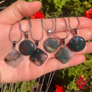 Puede incluir: Una colección de colgantes de piedras preciosas en cadenas de plata. Los colgantes varían en forma, incluyendo redondos y en forma de diamante, y presentan una mezcla de tonos verdes, marrones y rosas. Los collares se muestran sobre un fondo natural y frondoso.