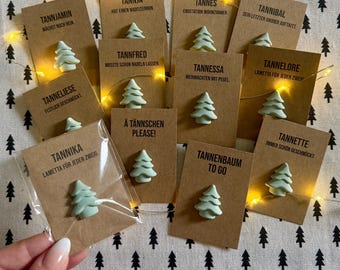 Op de afbeelding: Een verzameling lichtgroene kerstboomspeldjes bevestigd op individuele bruine kartonnen displaykaarten, elk met unieke Duitse namen en zinnen, gerangschikt op een stoffen ondergrond met een snoer warm licht.