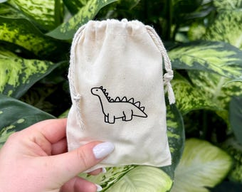 Op de afbeelding: Een klein, wit trekkoordtasje met een zwarte dinosaurus illustratie. De tas is gemaakt van natuurlijke stof en heeft een eenvoudig ontwerp.