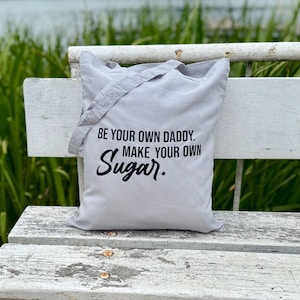 Könnte beinhalten: Hellgraue Tragetasche mit dem schwarzen Text "BE YOUR OWN DADDY. MAKE YOUR OWN Sugar.". Die Tasche hat einen Schulterriemen und besteht aus weichem Material. Sie liegt auf einer weißen Bank.