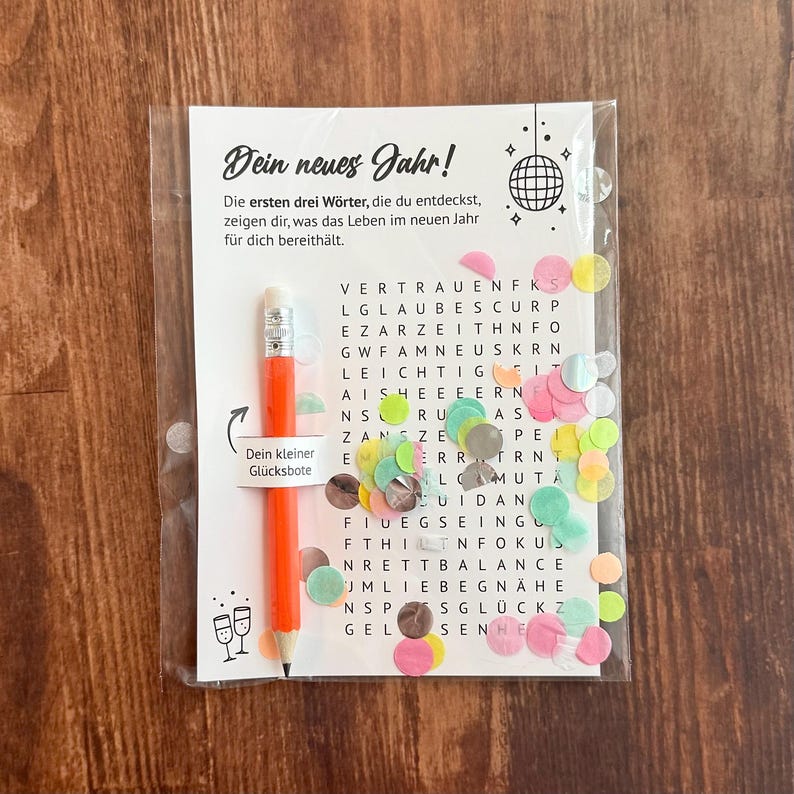 Op de afbeelding: Een doorzichtige plastic zak met een woordzoekpuzzel met de tekst "Dein neues Jahr!" en een oranje potlood. De puzzel bevat kleurrijke confetti en een potlood met de tekst "Dein kleiner Gl&uuml;cksbote".