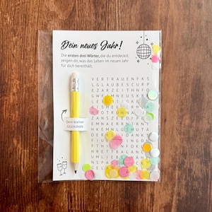 Op de afbeelding: Een doorzichtige plastic verpakking met een wenskaart en een gele potlood. De kaart heeft Duitse tekst en een woordzoekpuzzel met kleurrijke confetti. Het potlood heeft een zilveren punt en een gum.