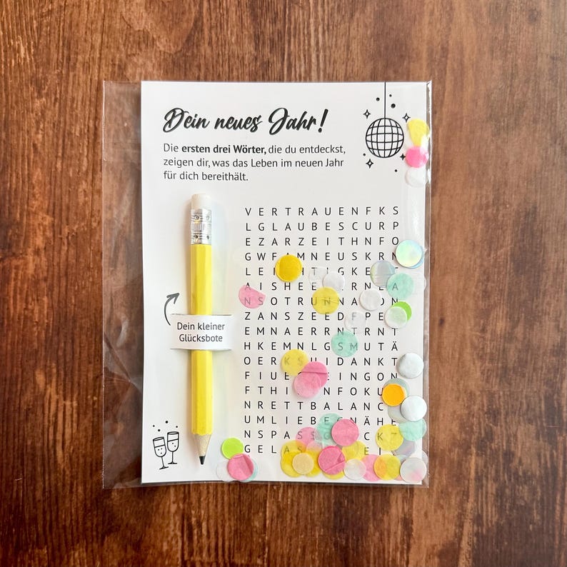 Op de afbeelding: Een doorzichtige plastic verpakking met een wenskaart en een gele potlood. De kaart heeft Duitse tekst en een woordzoekpuzzel met kleurrijke confetti. Het potlood heeft een zilveren punt en een gum.
