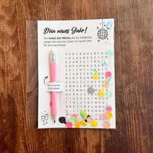 Op de afbeelding: Een roze potlood en een woordzoekkaart in een transparante plastic hoes. De kaart heeft de tekst "Dein neues Jahr!". Kleurrijke confetti en iriserende stickers zijn verspreid rond de kaart.