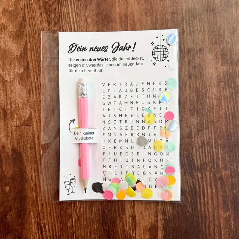 Op de afbeelding: Een roze potlood en een woordzoekkaart in een transparante plastic hoes. De kaart heeft de tekst "Dein neues Jahr!". Kleurrijke confetti en iriserende stickers zijn verspreid rond de kaart.