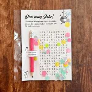 Op de afbeelding: Een cadeaupakket met Nieuwjaarsthema met een roze potlood, een woordzoekpuzzel en confetti. Het potlood heeft een label met de tekst "Je kleine geluksbrenger". De puzzel heeft Duitse tekst.
