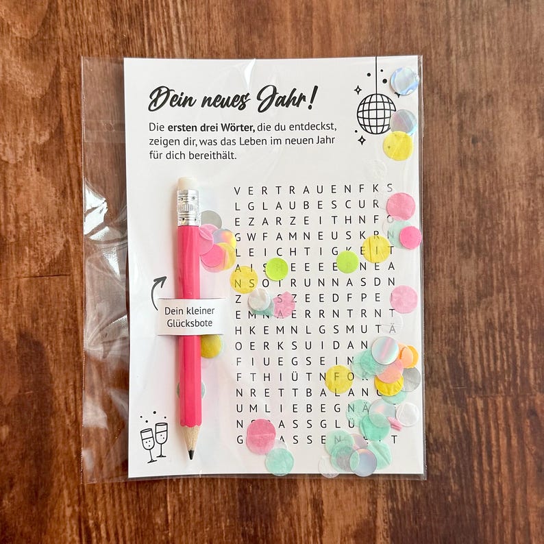 Op de afbeelding: Een cadeaupakket met Nieuwjaarsthema met een roze potlood, een woordzoekpuzzel en confetti. Het potlood heeft een label met de tekst "Je kleine geluksbrenger". De puzzel heeft Duitse tekst.