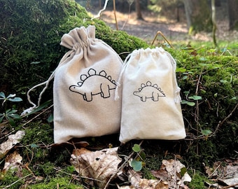 Op de afbeelding: Twee kleine, natuurlijke kleur stoffen zakken met trekkoord sluitingen. Elke zak heeft een zwarte lijn tekening van een dinosaurus met een stekelige rug.