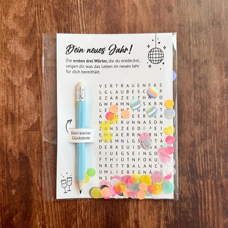 Op de afbeelding: Een doorzichtige plastic verpakking met een woordzoekpuzzel met de tekst "Dein neues Jahr!" en een lichtblauwe potlood. De puzzel is gevuld met letters en omgeven door kleurrijke confetti.