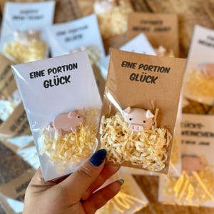 Könnte beinhalten: Ein kleines rosa Schweinchen in einer transparenten Plastiktüte, eingebettet in gelbes Papier. Die Tüte ist an einer weißen Karte mit dem deutschen Text "EINE PORTION GLÜCK" befestigt. Weitere ähnliche Pakete sind im Hintergrund zu sehen.