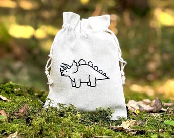 Op de afbeelding: Een witte trekkoordtas met een zwarte omtrek van een triceratops-dinosaurus. De tas ligt op een bed van groen mos.